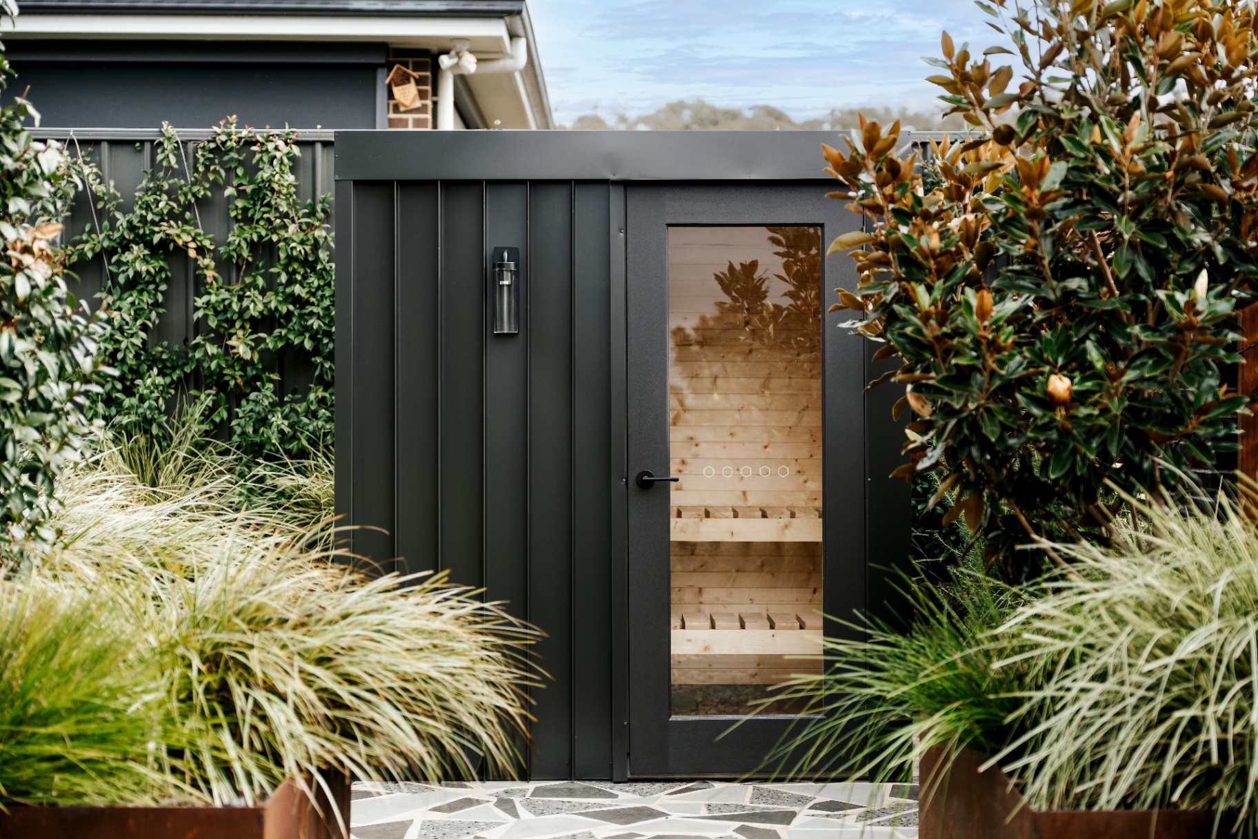 Black Colorbond sauna with glass door in a lush garden setting